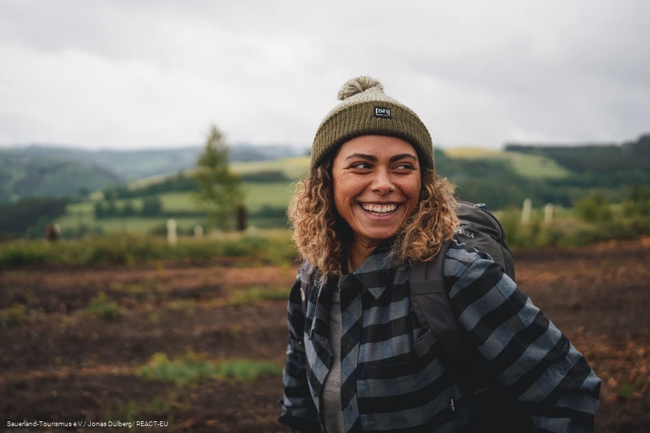 Eine Frau steht in Wanderausrüstung in einer idyllischen Landschaft und ist sichtlich glücklich