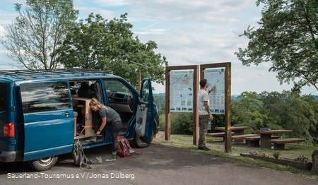 Zwei Wanderer vor einem Van bereiten sich auf eine Wanderung vor, im Hintergrund die Meinhardus Mattenschanze in Meinerzhagen