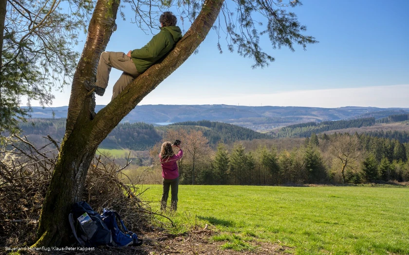 Ein Mann sitzt im Baum und genießt die Aussicht auf den Biggesee. Eine Frau macht im Hintergund ein Selfie am Sauerland-Höhenflug