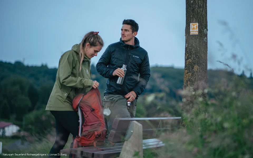 Zwei Wanderer  stehen an einer Bank am Sauerland-Höhenflug und packen ihren Rucksack