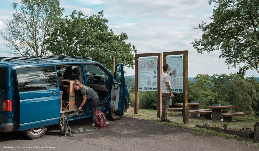 Zwei Wanderer vor einem Van bereiten sich auf eine Wanderung vor, im Hintergrund die Meinhardus Mattenschanze in Meinerzhagen