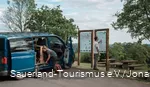 Zwei Wanderer vor einem Van bereiten sich auf eine Wanderung vor, im Hintergrund die Meinhardus Mattenschanze in Meinerzhagen
