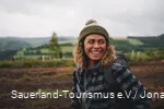Eine Frau steht in Wanderausrüstung in einer idyllischen Landschaft und ist sichtlich glücklich